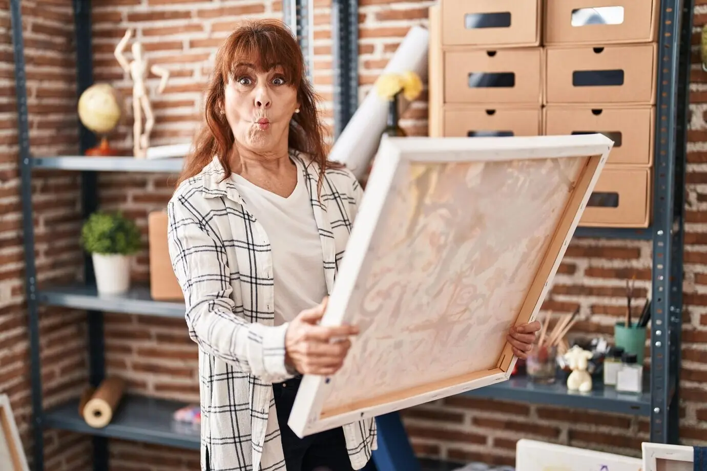 Eine mittelalte hispanische Frau blickt auf eine Malerleinwand, zieht mit dem Mund eine Fischgrimasse und kneift die Augen zusammen – verrückt und komisch.